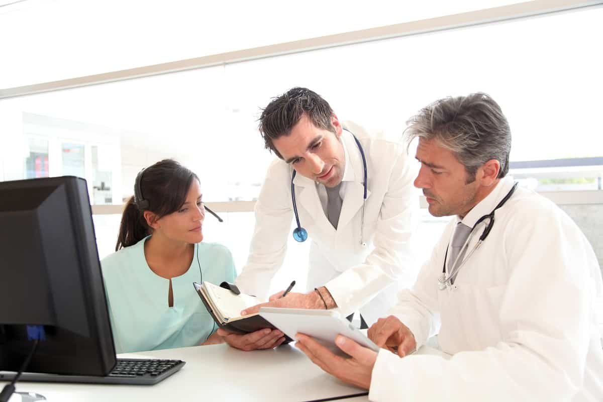 Female in a green top, with a man in a white coat, and another man standing between them looking at patient files.