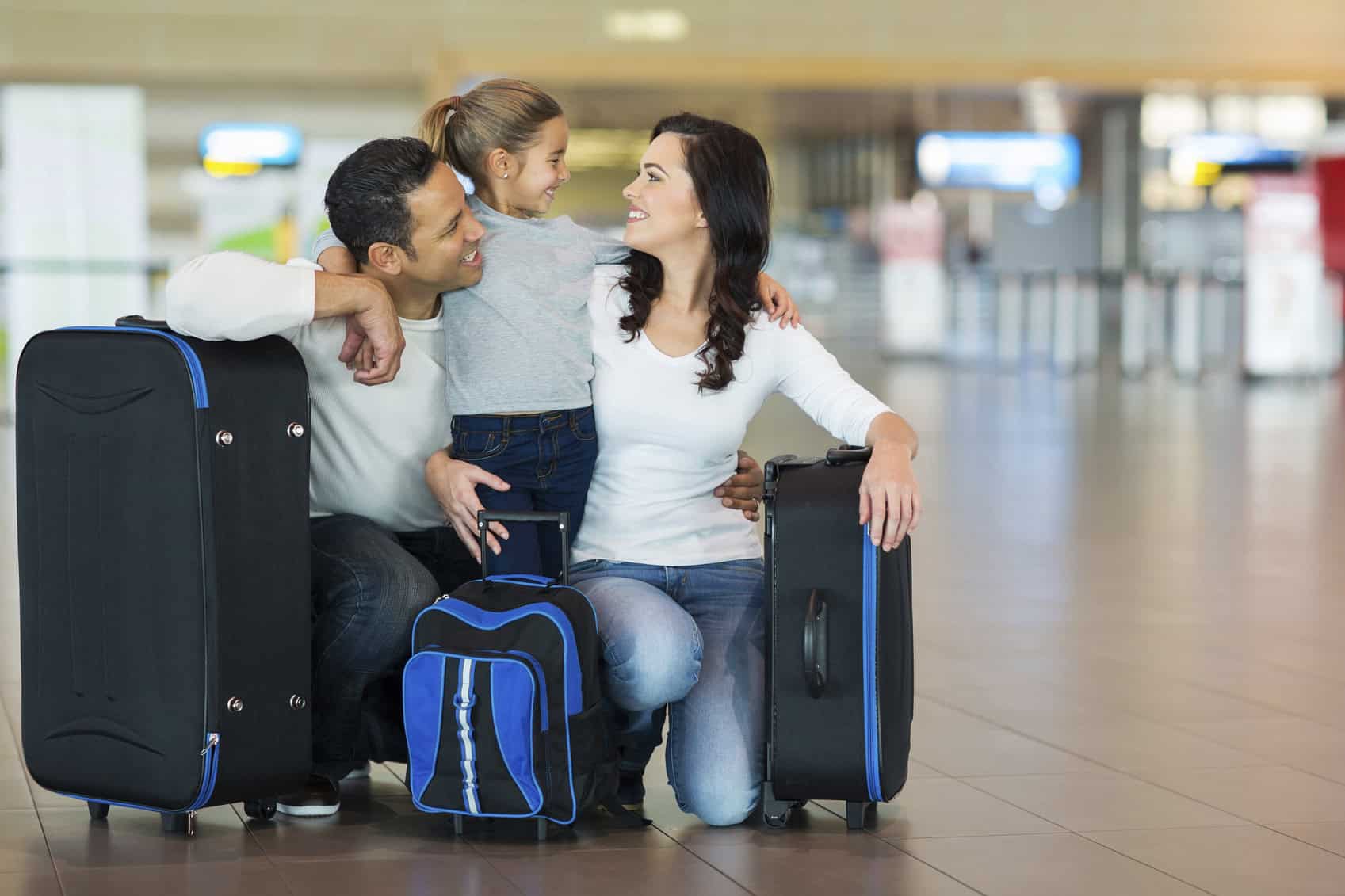 With an airport as the background, a smiling father and mother kneel to embrace their short daughter with three suitcases around them.