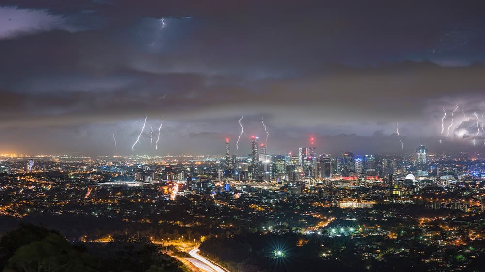 Heavy black clouds are lit by numerous lightning strikes against a city backdrop with many highrises.