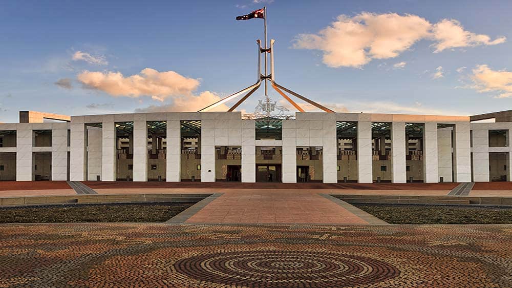 The front of Australia's Federal Parliament House with Aboriginal mosaic tiling in the front and the flag flying above it against blue skies and clouds.