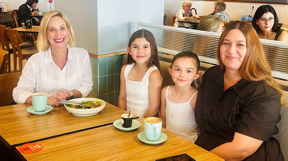 Justine Cain, Olivia with straight, long brown hair parted in the middle, Emily, with her hair pulled off her face in a tight ponytail, and their Mum, Yvette, sit at a booth at a coffee shop with their drinks and food on a wooden table.