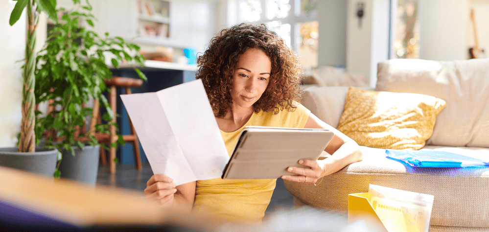 Woman with light brown curly hair to her shoulders holds a sheet of paper in one hand and compares the information on the tablet she is holding while sitting on a sofa.