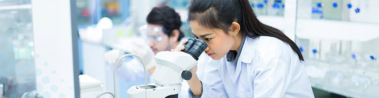 A young female researcher is looking into a double-lens microscope.